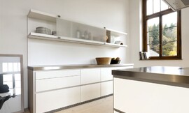 Deze moderne witte designkeuken maakt met zijn roestvrijstalen aanrechtbladen en greeploze witte fronten een strakke indruk. Aan de wand is een keukenblok geplaatst met grote, praktische lades. De lage kast daarboven heeft verschuifbare matglazen fronten en is voor een deel open. Het kookeiland heeft hetzelfde design: roestvrijstalen werkblad, witte greeploze fronten. Niets verstoort hier de kalmte die deze keuken uitstraalt. Zuordnung: Stil Design-keukens, Planungsart Keuken met keukeneiland