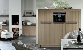 Hier ziet u een detailfoto van de hoogkast/apparatuurkast met brede wangen, stanggrepen en verticale strepen in het hout. Ook ziet u hoe hetzelfde concept is doorgevoerd in de woonkamer. Aparte eettafel met rieten stoelen. Zuordnung: Stil Landelijke keukens, Planungsart Binneninrichting van de keuken