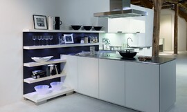 Deze open keuken heeft een gereduceerde look met witte fronten en een dun, metallic aanrechtblad. Het kookeiland staat haaks op een open wandgedeelte. Deze open wand met legplanken is ideaal om alles direct bij de hand te hebben, maar kan tegelijkertijd ook decoratief worden gebruikt. De blauwe achtergrond van de open kast zorgt voor een speels accent. Op deze open wandkast sluit het keukenblok met de spoelbak aan. De onder- en bovenkasten bieden voldoende bergruimte. Een keukenplanning zoals deze is ook erg geschikt voor grote ruimtes die deels als woonkamer functioneren. Zuordnung: Stil Luxe keukens, Planungsart Open keuken (woonkeuken)